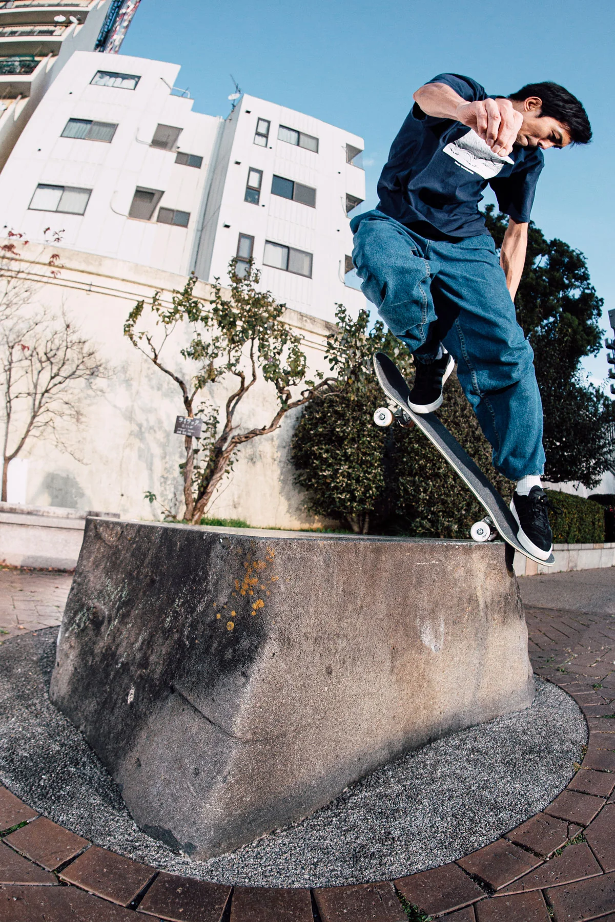 A low-angle action shot of a skateboarder Shin Sanbongi balancing mid-back noseblunt slide on the top edge of a large concrete block with white apartment buildings in the background.