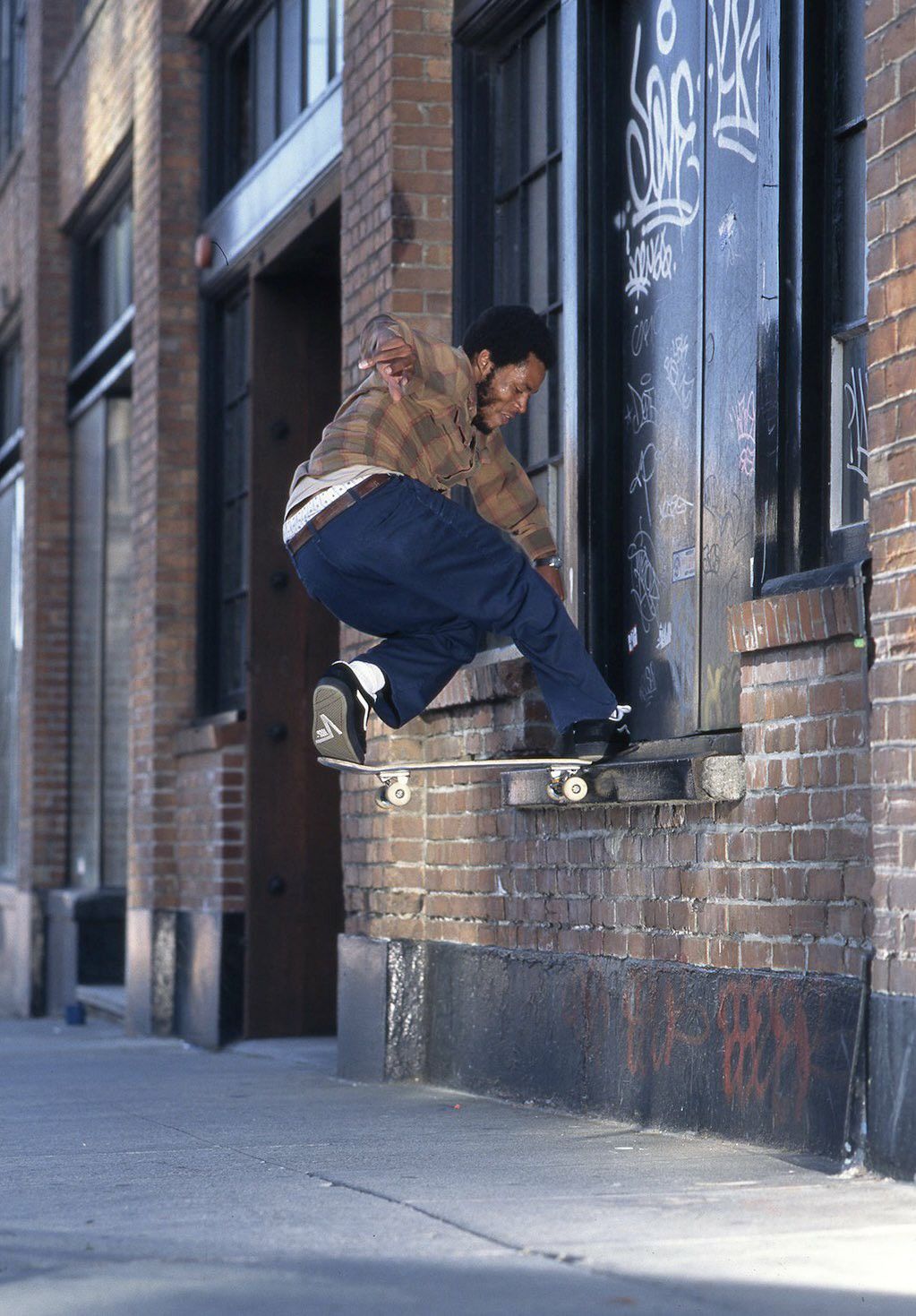 A street photograph of skateboarder Ray Barbee in a plaid shirt doing a frontside noseslide on a ledge along a city sidewalk.