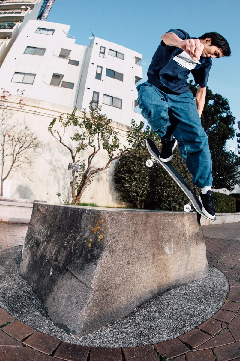 A low-angle action shot of a skateboarder Shin Sanbongi balancing mid-back noseblunt slide on the top edge of a large concrete block with white apartment buildings in the background.